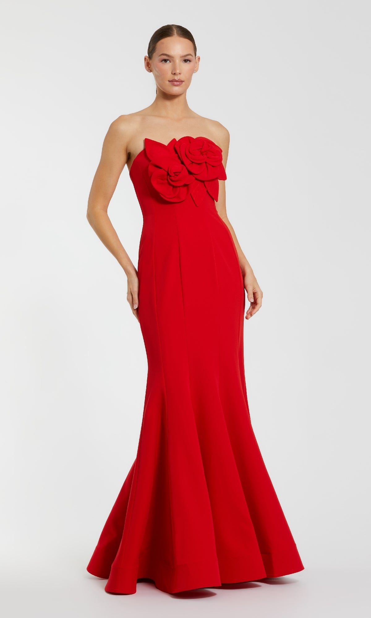A woman wears the Mac Duggal 2300 Strapless Long Black Formal Dress, featuring two large fabric roses on the bodice, standing against a plain light background.